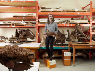 David Evans stands on a box in a warehouse surrounded by fossils.